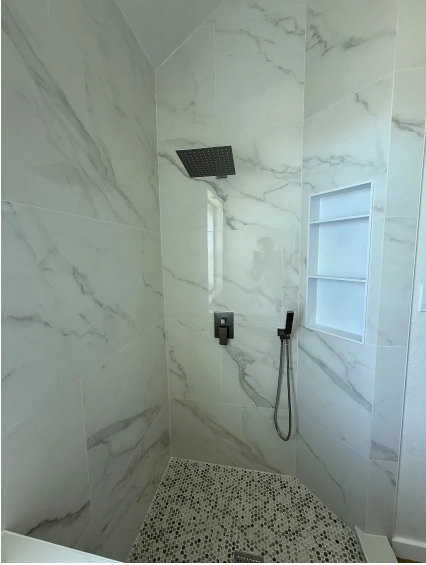 Modern walk-in shower with large white marble-like tiles, a black square rainfall shower head, a handheld shower wand, a built-in niche with shelves, and pebble stone flooring.