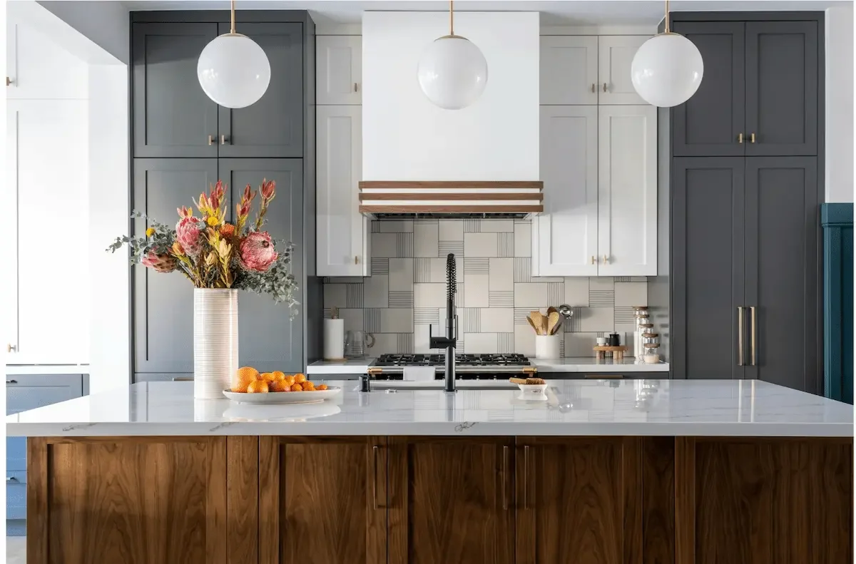 Modern kitchen with a white marble island, dark blue upper cabinets, white lower cabinets, and a patterned backsplash. There is a vase of flowers and a plate of oranges on the island, with three spherical pendant lights hanging above.