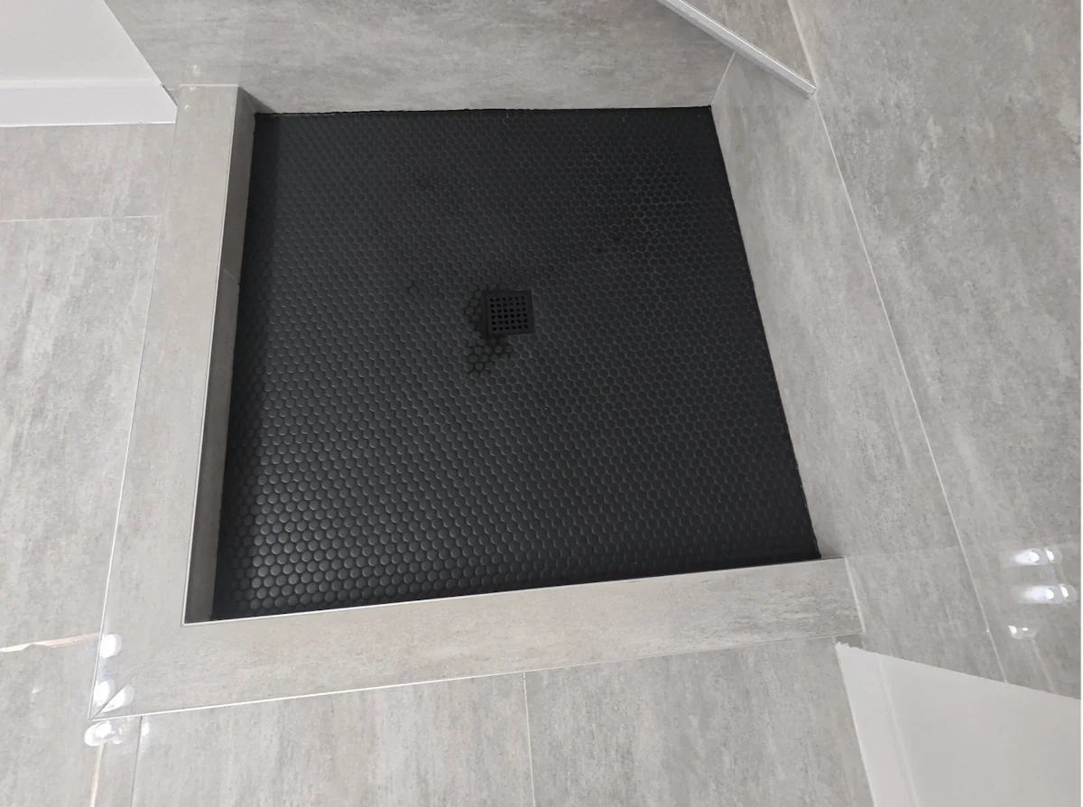 Shower drain in a black, textured shower floor with gray tiled walls.