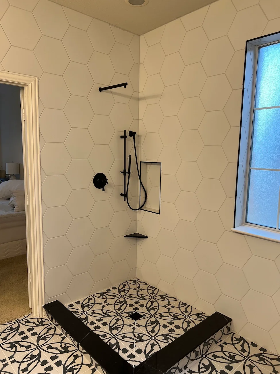 Modern walk-in shower with black fixtures, geometric black and white floor tiles, and white hexagon wall tiles, next to a frosted window.