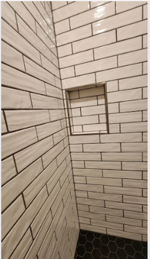 Interior view of a tiled shower with white subway tiles and a black hexagonal tile floor, featuring a built-in shelf in the wall.