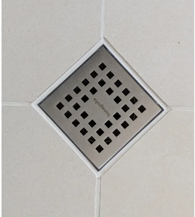 Square metal floor drain with small black square holes, installed in a tiled white floor.