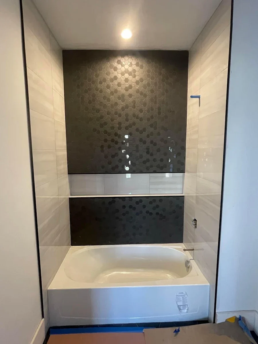 Bathroom with beige tiled walls, a black decorative tile panel, a white bathtub, and a ceiling light.