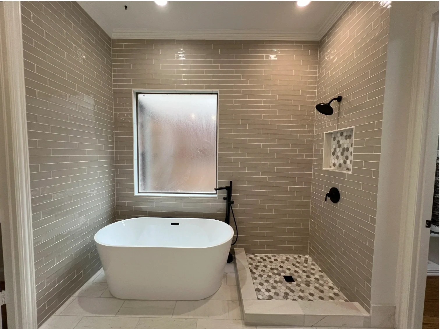 Modern bathroom with a freestanding bathtub, gray tiled walls, a frosted window, and a tiled walk-in shower with black fixtures and a small mosaic tile niche.