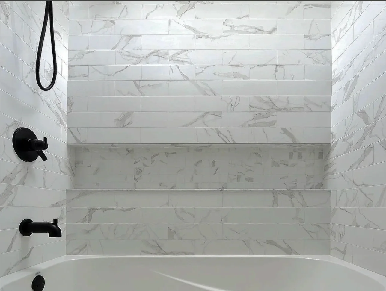 View of a modern shower with white marble tiles with gray veining on the walls, black fixtures, and a white bathtub at the bottom.