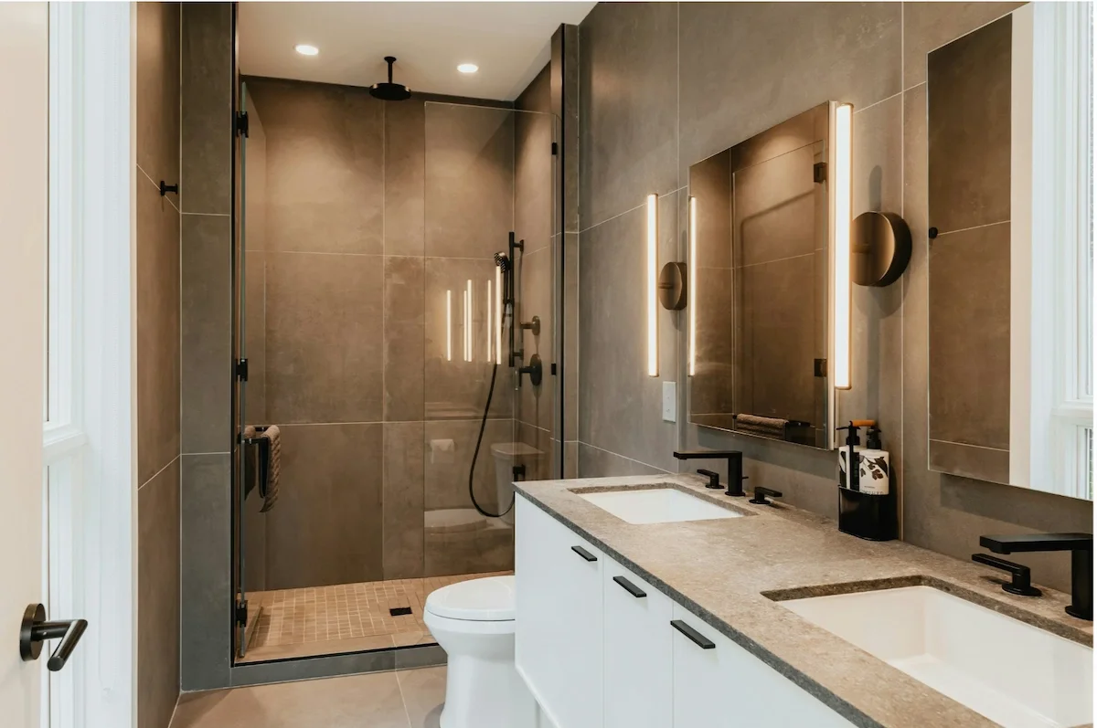 Modern bathroom with dual sinks, large mirror, black fixtures, and a shower area with a glass door and beige tiled walls.