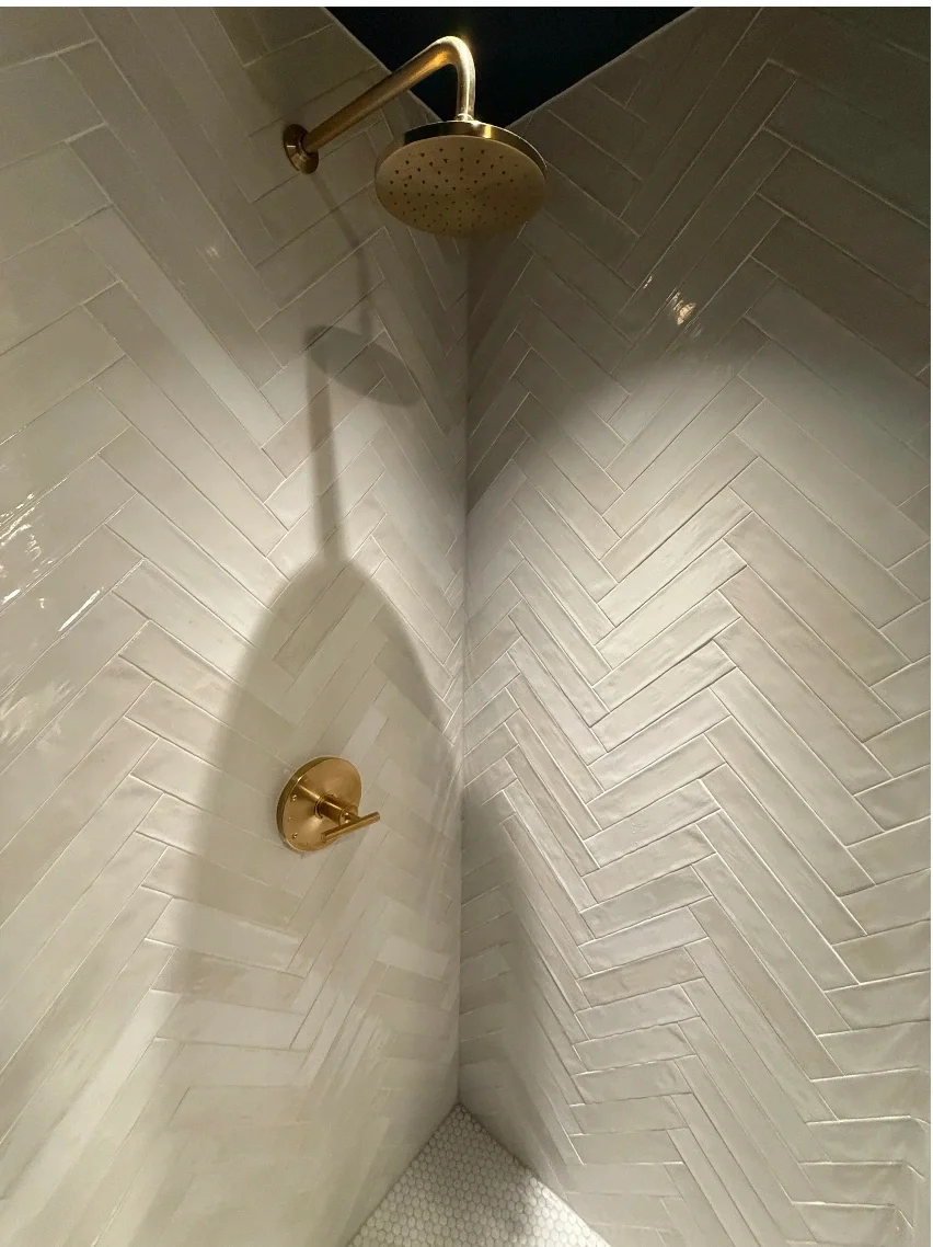A modern shower with beige herringbone tile walls, a brass showerhead, and a matching brass control handle.