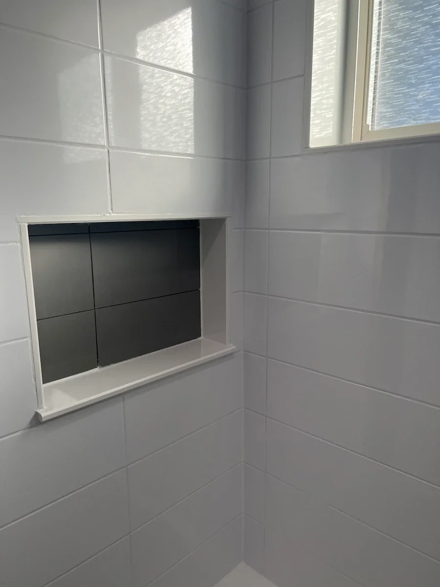 A close-up view of a tiled shower or bathroom wall with white tiles, a recessed shelf with black tiles inside, and a window with textured glass.