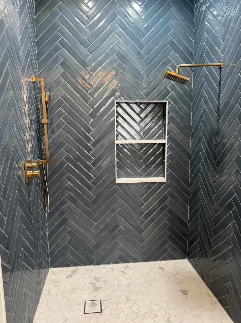 Shower with dark gray herringbone tile walls, a window frame on the wall, and white hexagon tile floor with a drain.