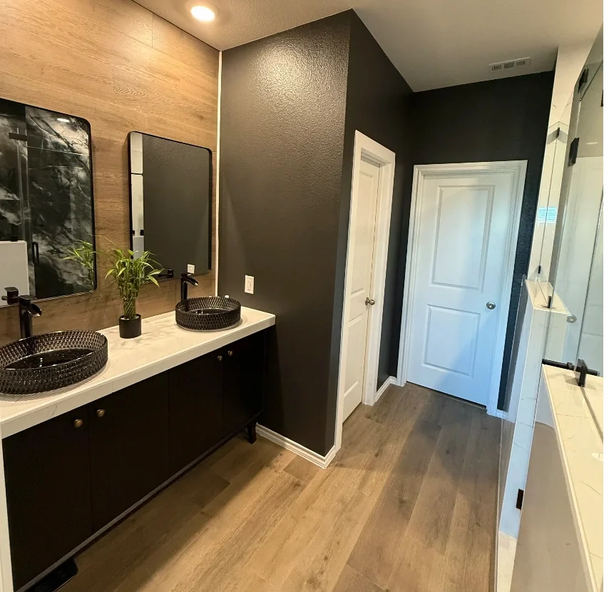 Modern bathroom with dual black vessel sinks on a white marble countertop, two black framed mirrors, a small green plant between the sinks, dark walls, and wooden accents.