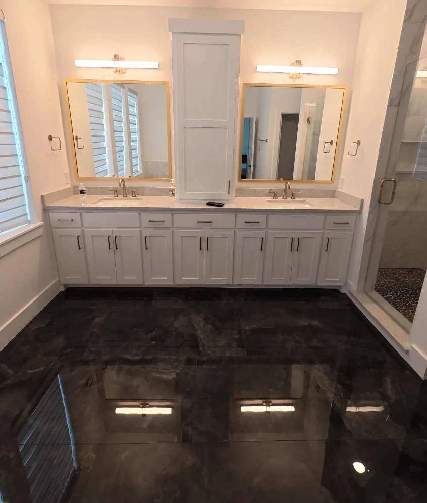Modern bathroom with dual sinks, large mirrors with gold trim, white cabinets, marble countertops, and a glass shower enclosure on the right. Shiny black marble floor and white walls with horizontal blinds on window.