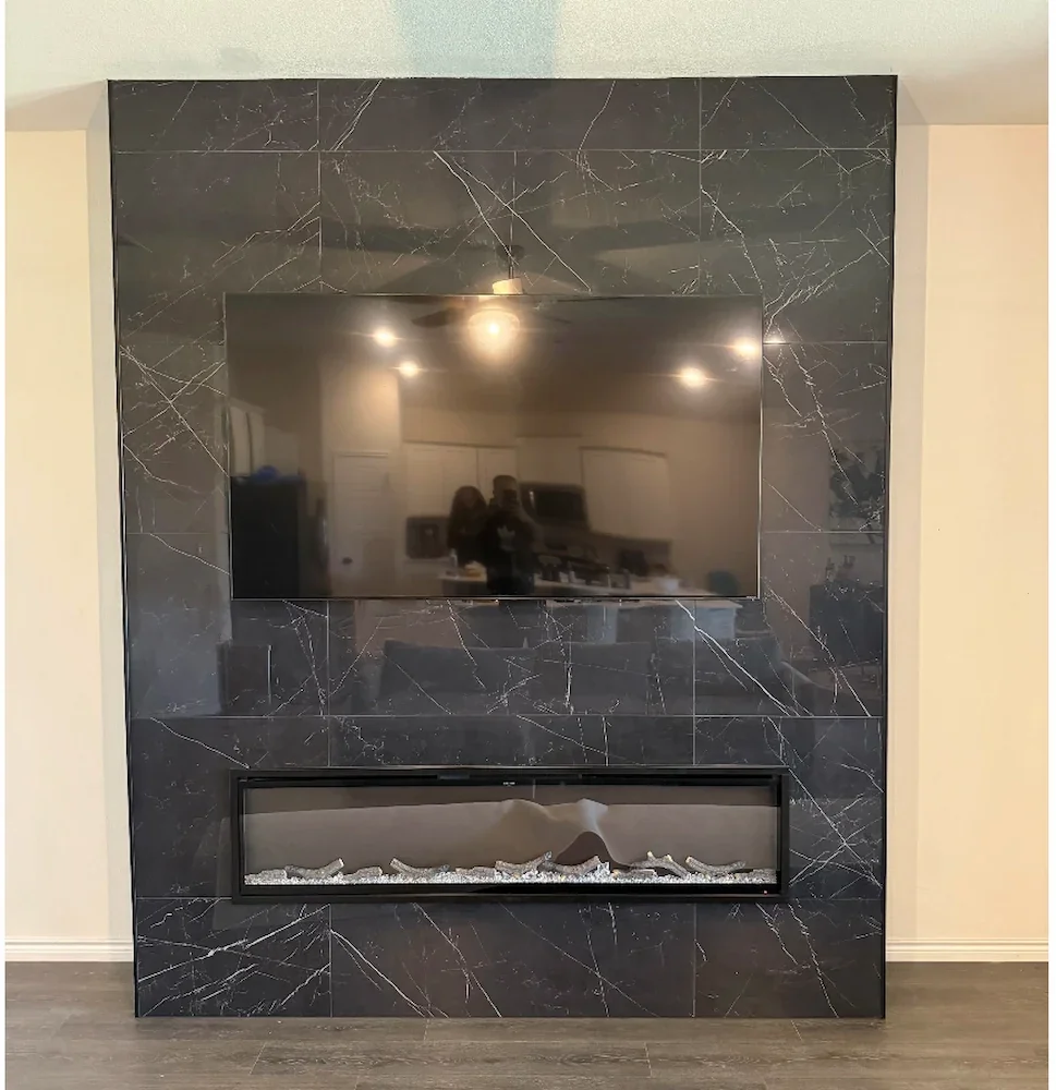 A black marble wall with white veining, featuring a mounted flat-screen TV and a built-in linear electric fireplace below it with decorative white stones.