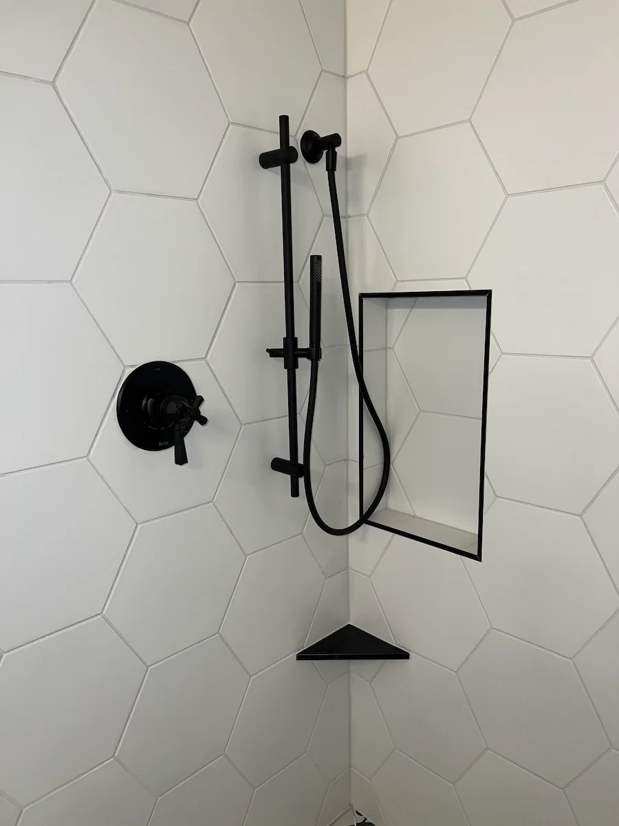 Black shower fixtures and a shower niche with a triangular shelf on a white hexagonal tiled shower wall.