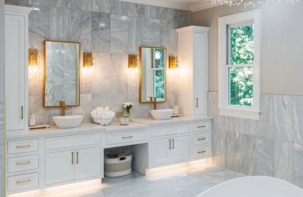 Modern bathroom with white cabinetry, marble walls and floors, two white vessel sinks, gold fixtures, two mirrors, and a window showing green trees outside.