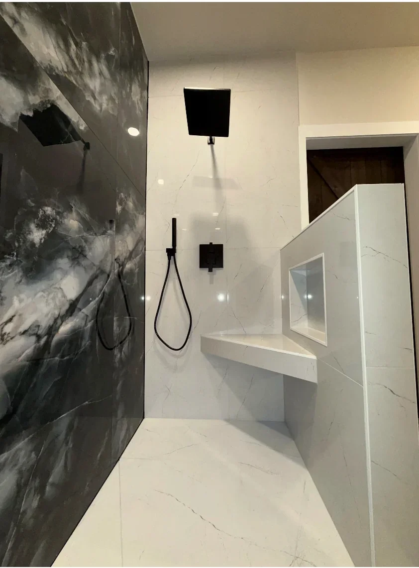 Modern walk-in shower with black rainfall showerhead, handheld shower, black fixtures, black marble wall, white marble tiles, built-in bench, and a white partition.
