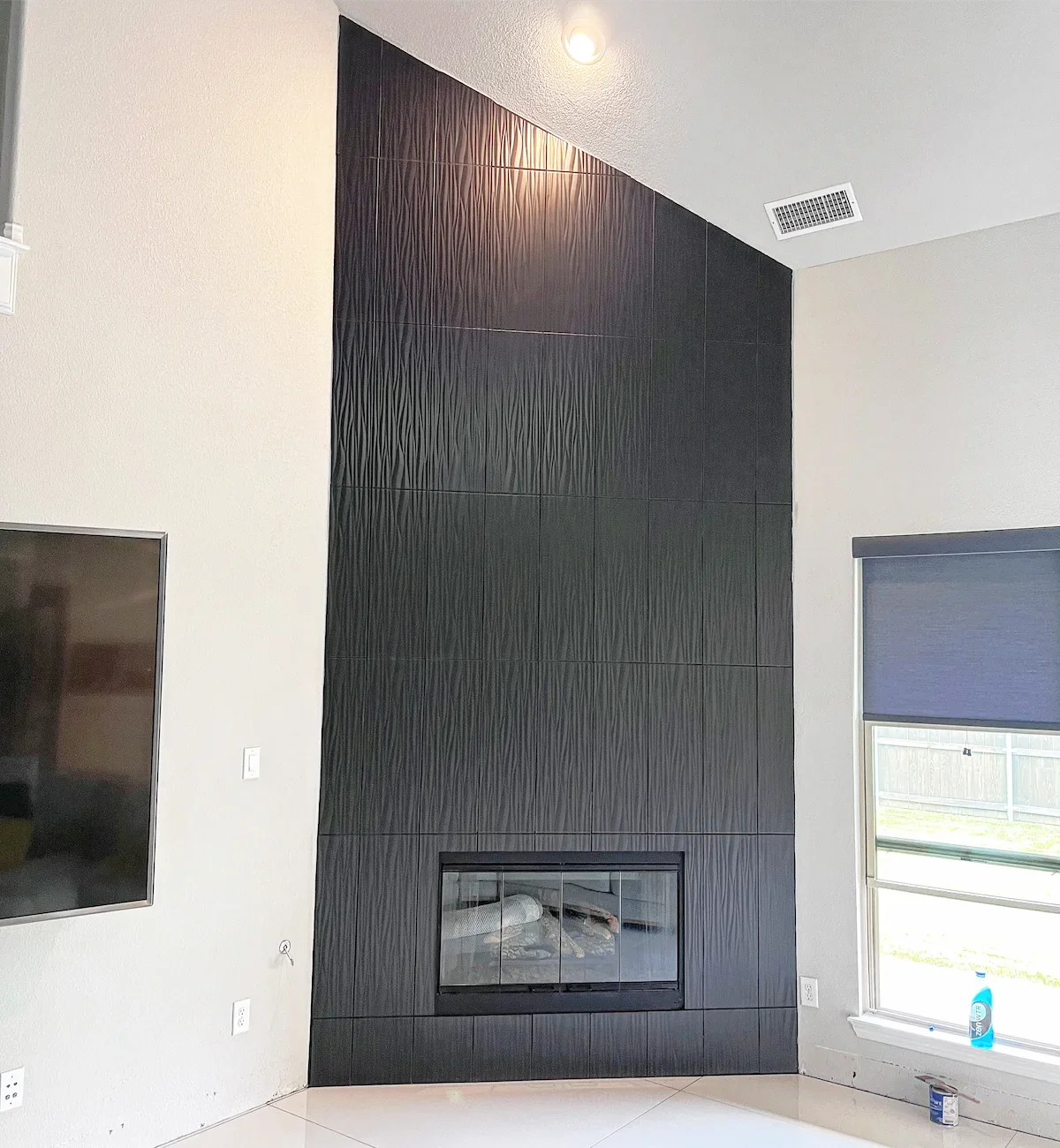 Modern black tiled fireplace on a light-colored wall in a living room with a large window to the right, with a blue bottle and a small can on the windowsill.