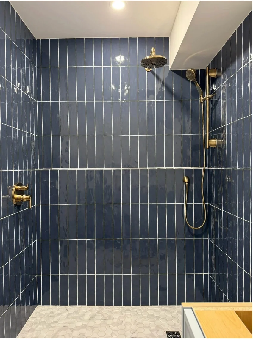 Modern shower with navy blue tiled walls, gold fixtures, a fixed showerhead, a handheld shower, and a small wooden bench in the corner.