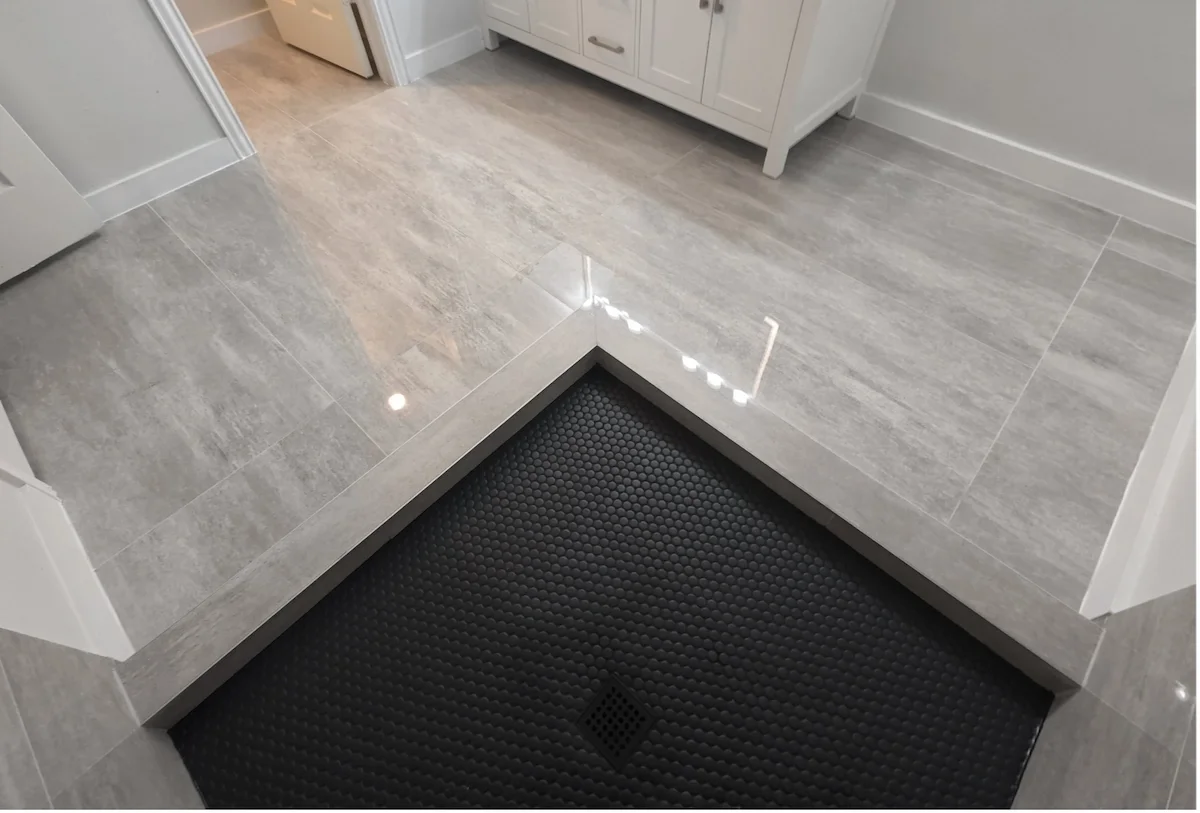 A bathroom or laundry room with gray tiled floor, white cabinetry, and a black rubber mat with a drain in front of a shower or wet area.