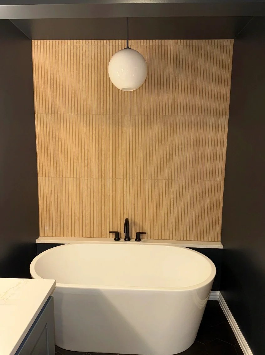 Modern bathroom with a white oval bathtub, black faucet, and a wooden slat feature wall, with a hanging round white light fixture above.