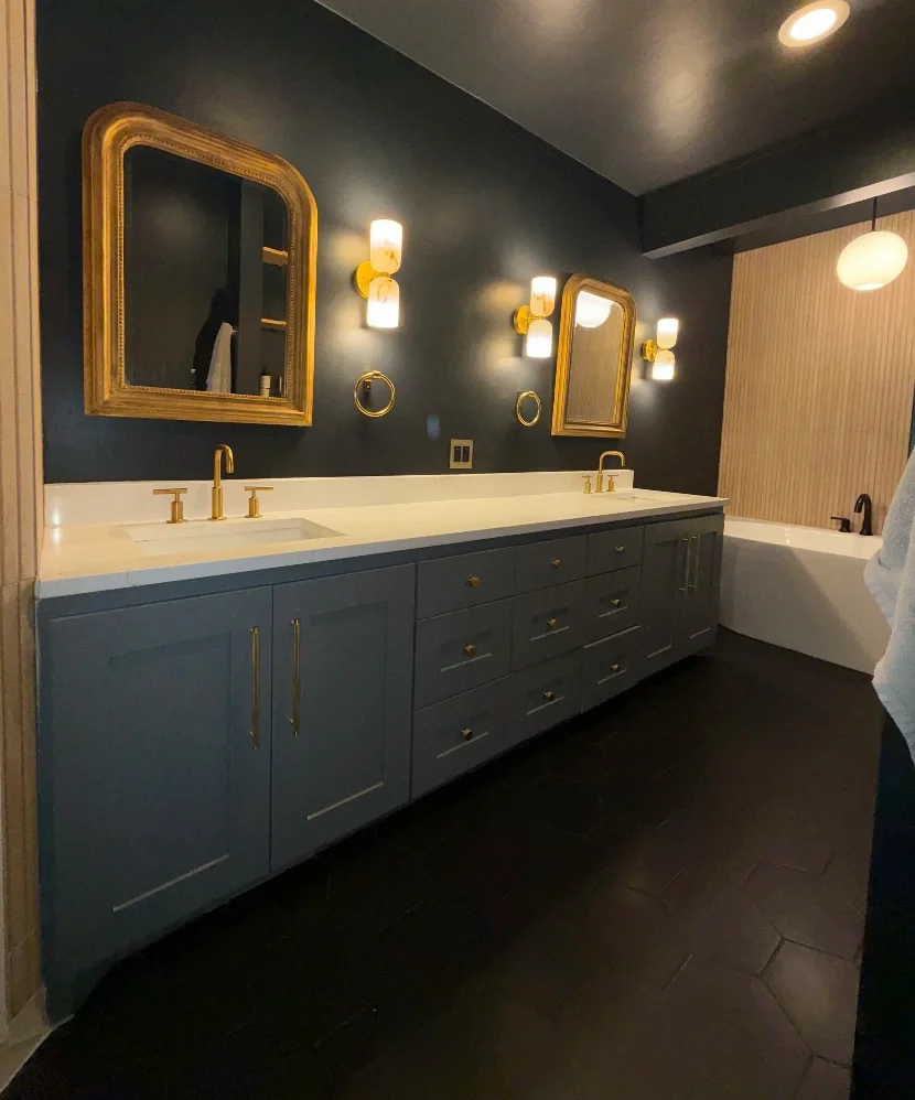 A modern bathroom with a dark blue vanity with gold hardware, two mirrors with gold frames, gold light fixtures, a white countertop with two sinks and gold faucets, a bathtub in the corner, and dark flooring.
