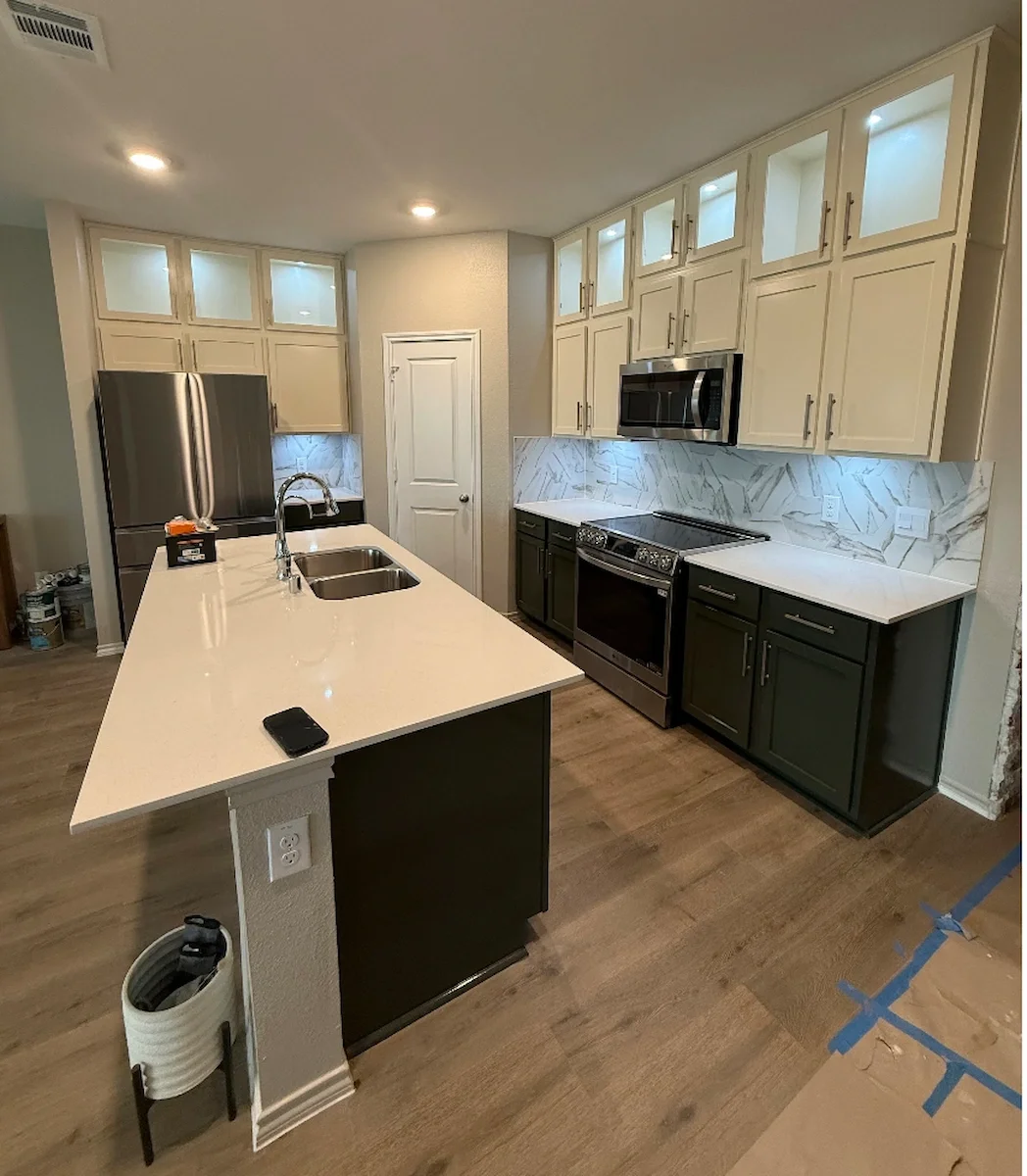 Modern kitchen with white upper cabinets, black lower cabinets, a white island with a double sink, stainless steel refrigerator, stove, and microwave, marble backsplash, and wooden flooring.