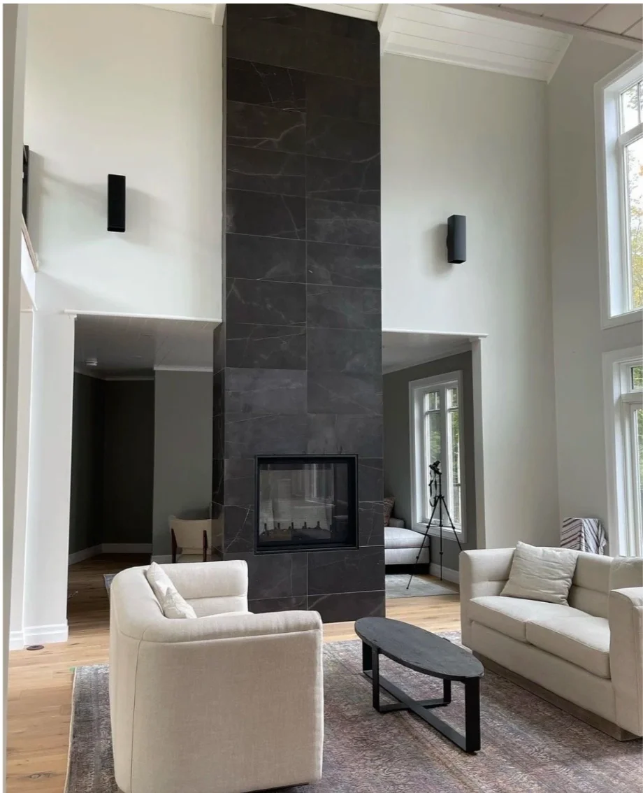 Living room with a large dark gray fireplace surrounded by white walls, featuring two white upholstered armchairs and a sofa, a black coffee table, and tall windows letting in natural light. A telescope is near the windows in the background.
