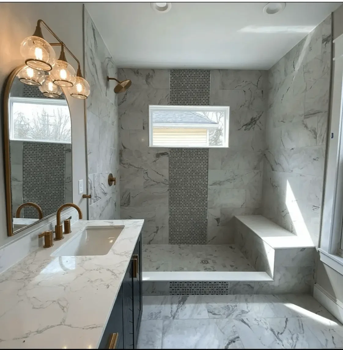 Modern bathroom with marble walls and floor, a built-in shower area with a bench, small window, gold fixtures, and a marble countertop with sink.