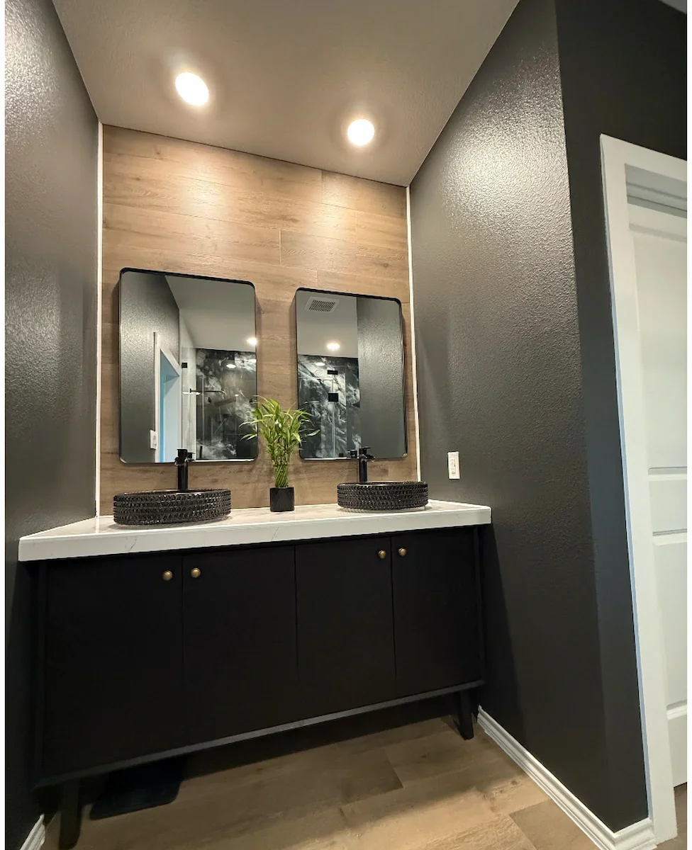 Modern bathroom vanity with two black vessel sinks, rectangular mirrors, a small plant in the center, dark wall paint, and wood flooring.