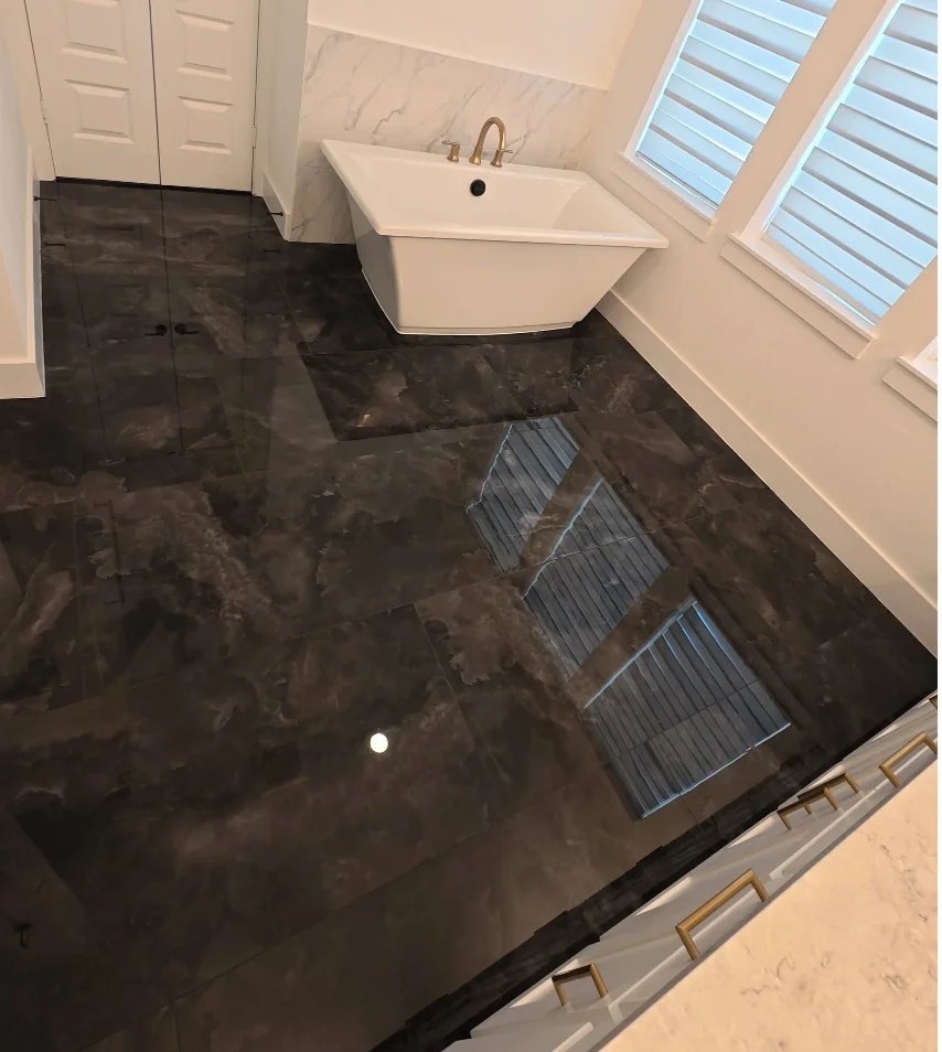 Bathroom featuring a white freestanding bathtub with gold fixtures, dark marbled tile floor, white walls with marble accents, and large windows with white blinds.