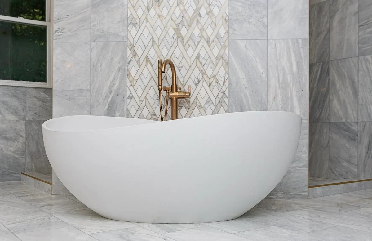 Modern white freestanding bathtub with gold faucet in a luxurious marble bathroom with a window.