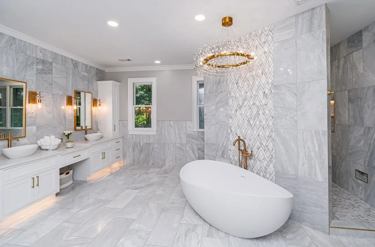 Luxurious modern bathroom with bathtub, double vanity, marble walls and floors, gold fixtures, and a decorative chandelier.