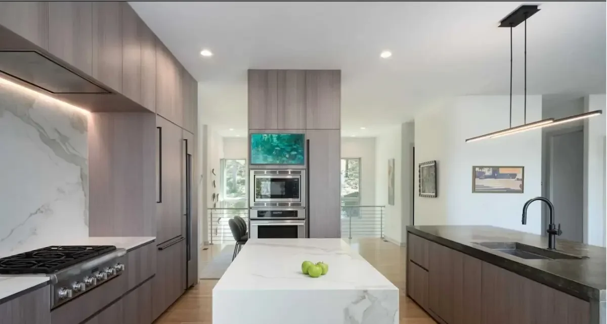 Modern kitchen with gray cabinetry, marble backsplash, and a large marble island with three green apples. To the right, there is a black sink with a black faucet, pendant lighting, and a dining area with a chair. In the background, built-in stainless