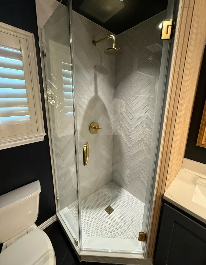 A glass shower enclosure with brass hardware in a bathroom, featuring white herringbone wall tiles and a small white hexagonal tile floor.