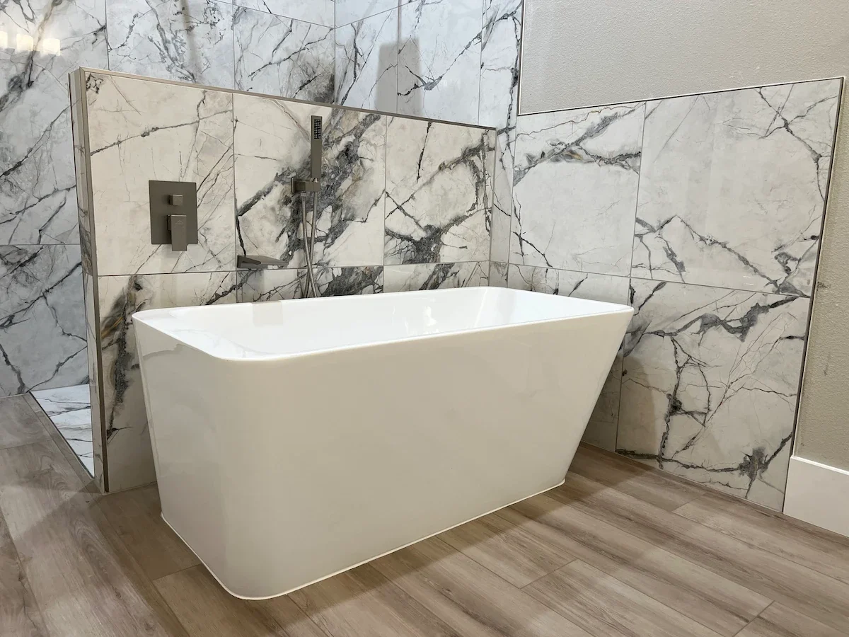 White freestanding bathtub with modern wall-mounted faucet and handheld showerhead in a bathroom with marble-tiled walls and wooden flooring.