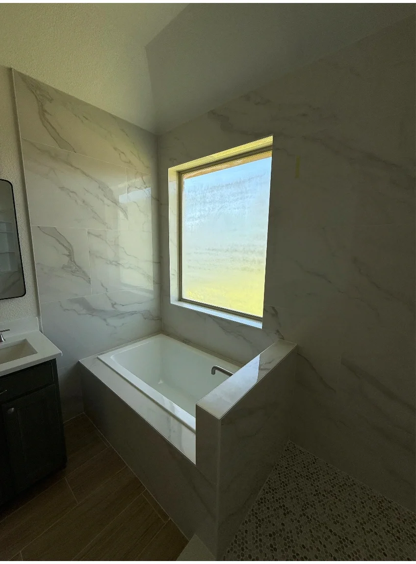 Bathroom with marble walls, a rectangular bathtub beneath a window, a small sink, and a tiled floor.