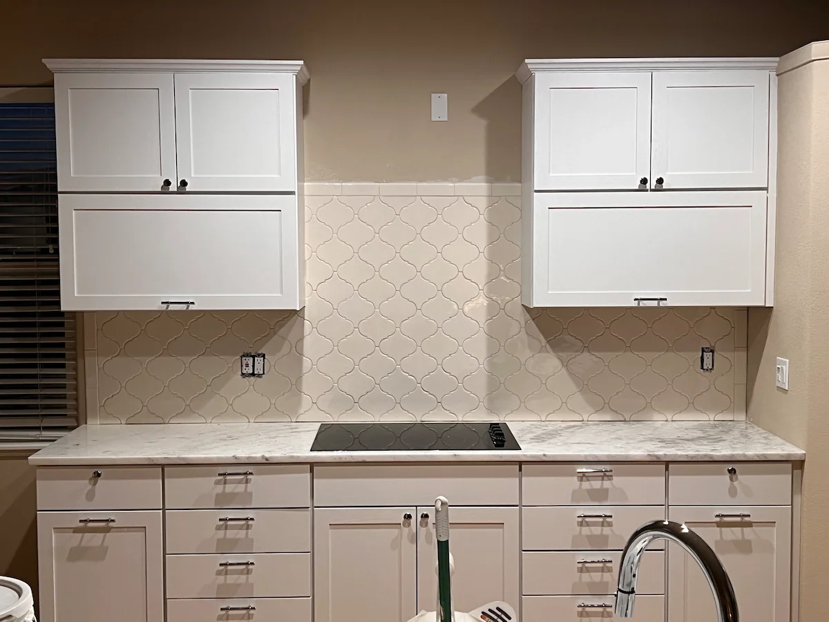 Kitchen with white cabinets, marble countertop, and beige patterned backsplash, with a black cooktop and a stainless steel faucet.