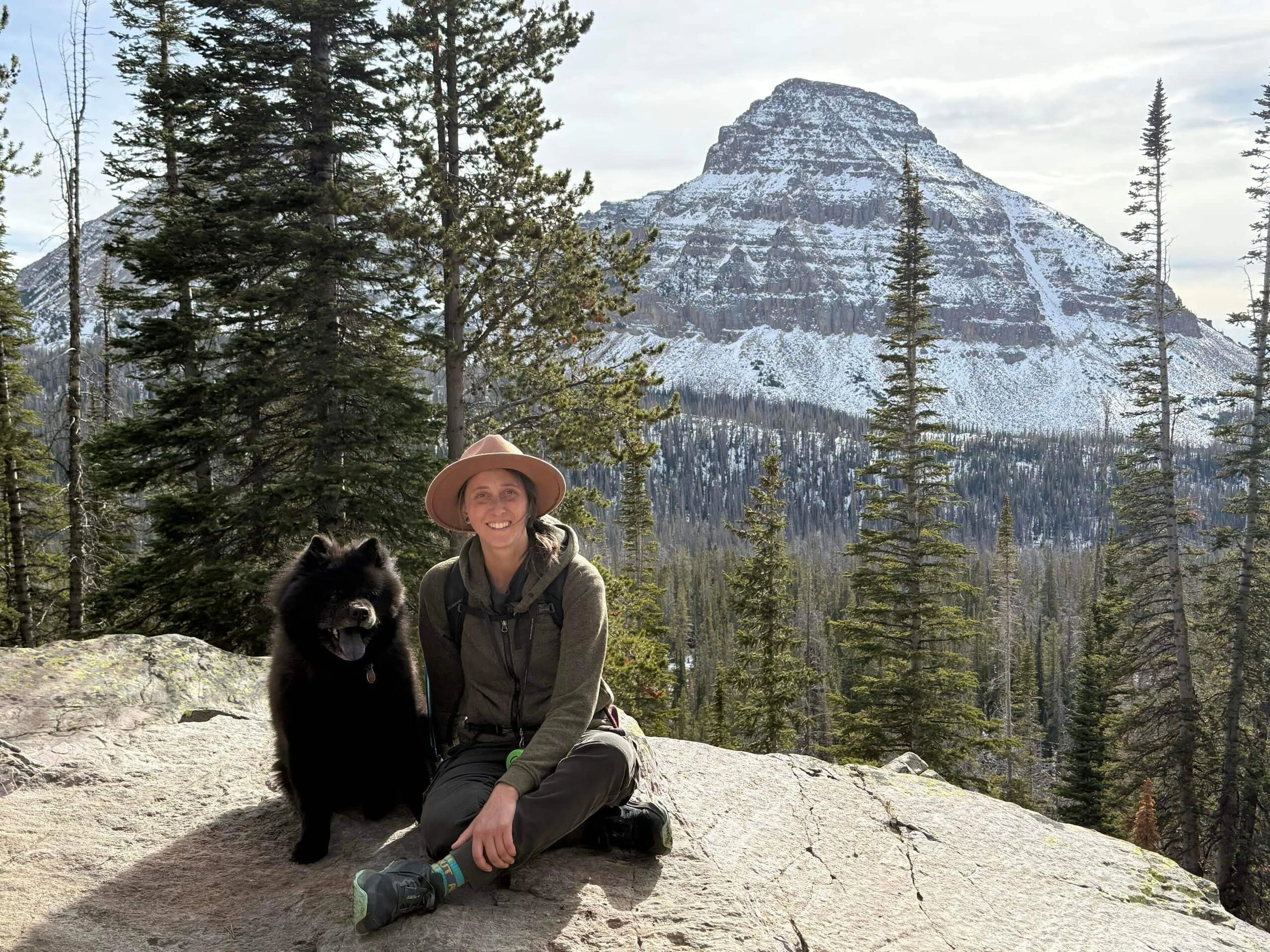 A woman and a black dog sitting on a large rock in a forest with tall pine trees and snow-covered mountain in the background.