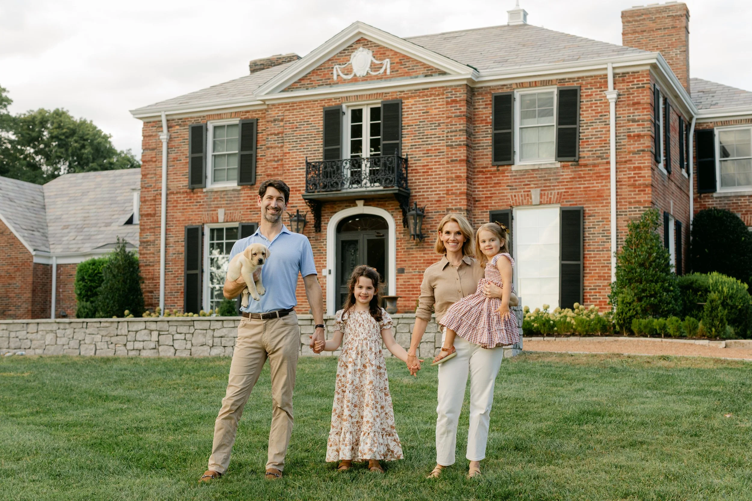 A family of four and a puppy standing in front of a large brick house with black shutters and a small balcony. The man is holding the puppy, the woman and girl are holding hands, and the woman is holding the little girl.