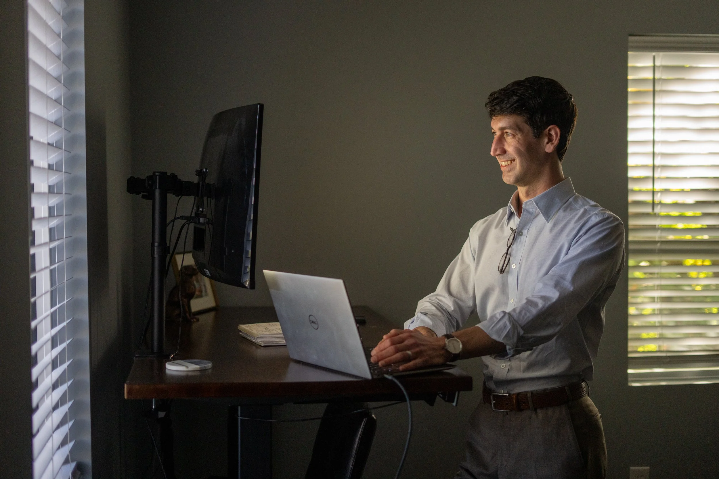 A man in a white shirt stands in front of a desk with a laptop and a monitor, smiling while working in a dimly lit room with closed window blinds.