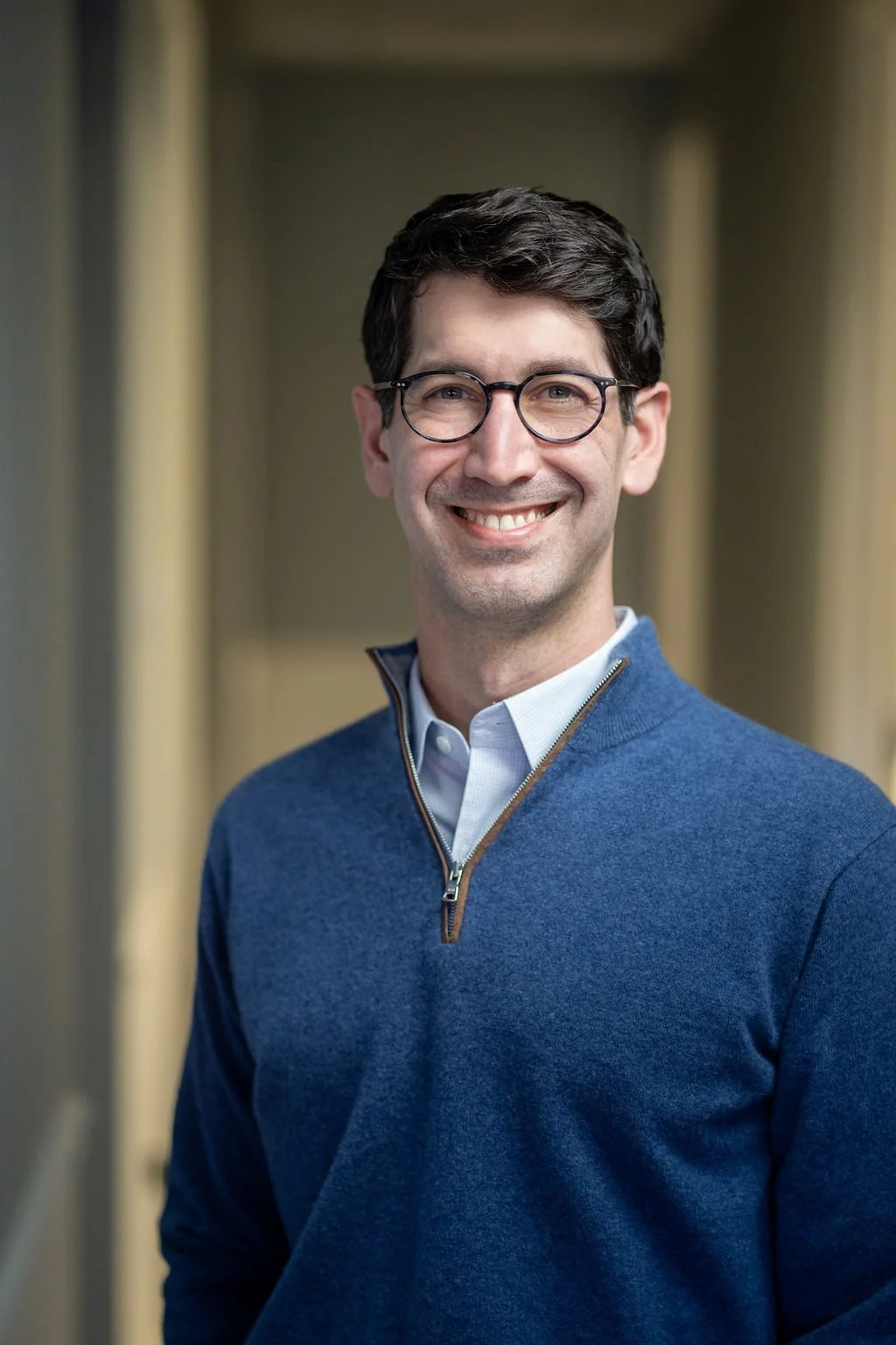A smiling man with dark hair, glasses, wearing a blue zip-up sweater over a collared shirt, standing in a corridor.