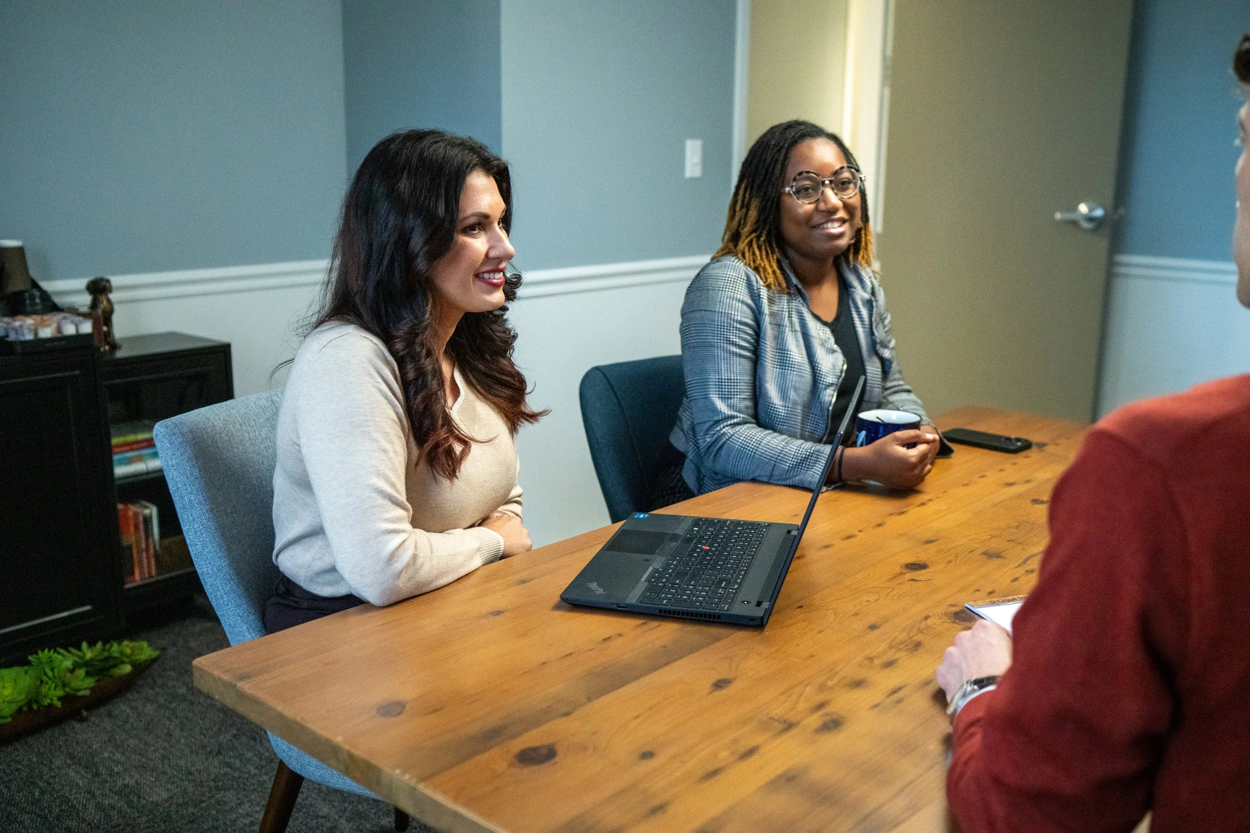 Two women sitting at a wooden table, engaging in a conversation with a person whose back is toward the camera, in an office setting. A laptop and a coffee mug are on the table.