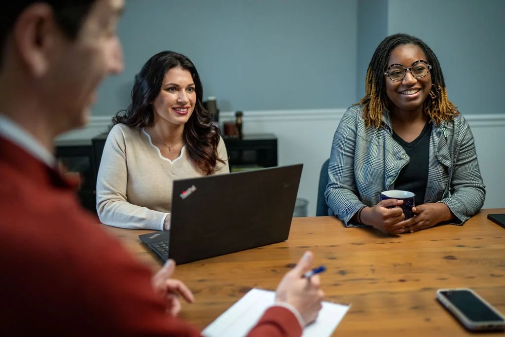 Three women sitting around a wooden table during a meeting, with a laptop, notebook, pen, and mug visible.