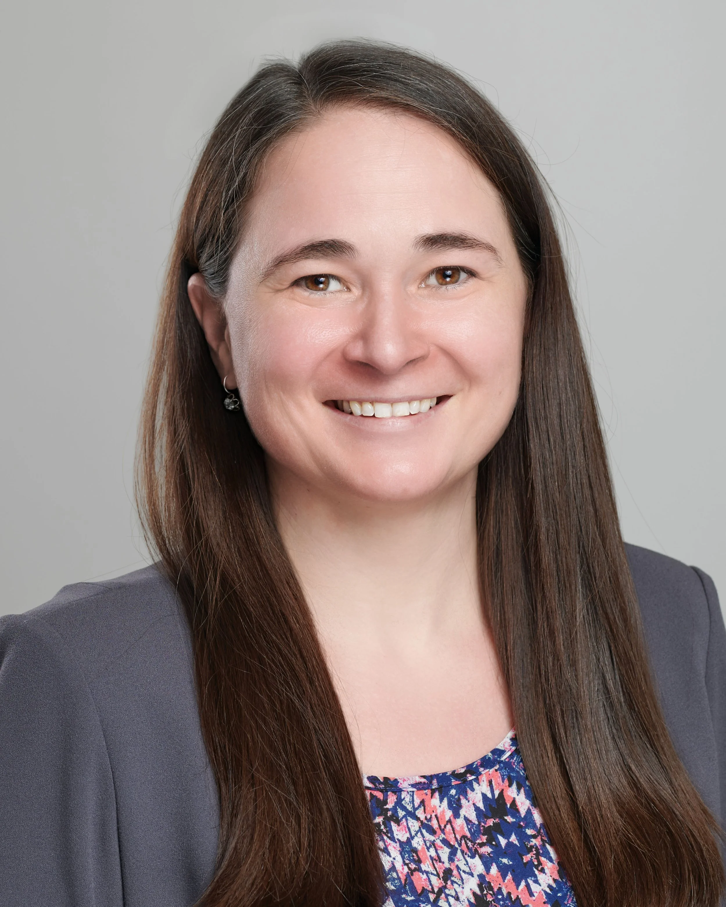 Headshot of a smiling woman with long brown hair, wearing a dark blazer and patterned blouse, against a plain gray background.