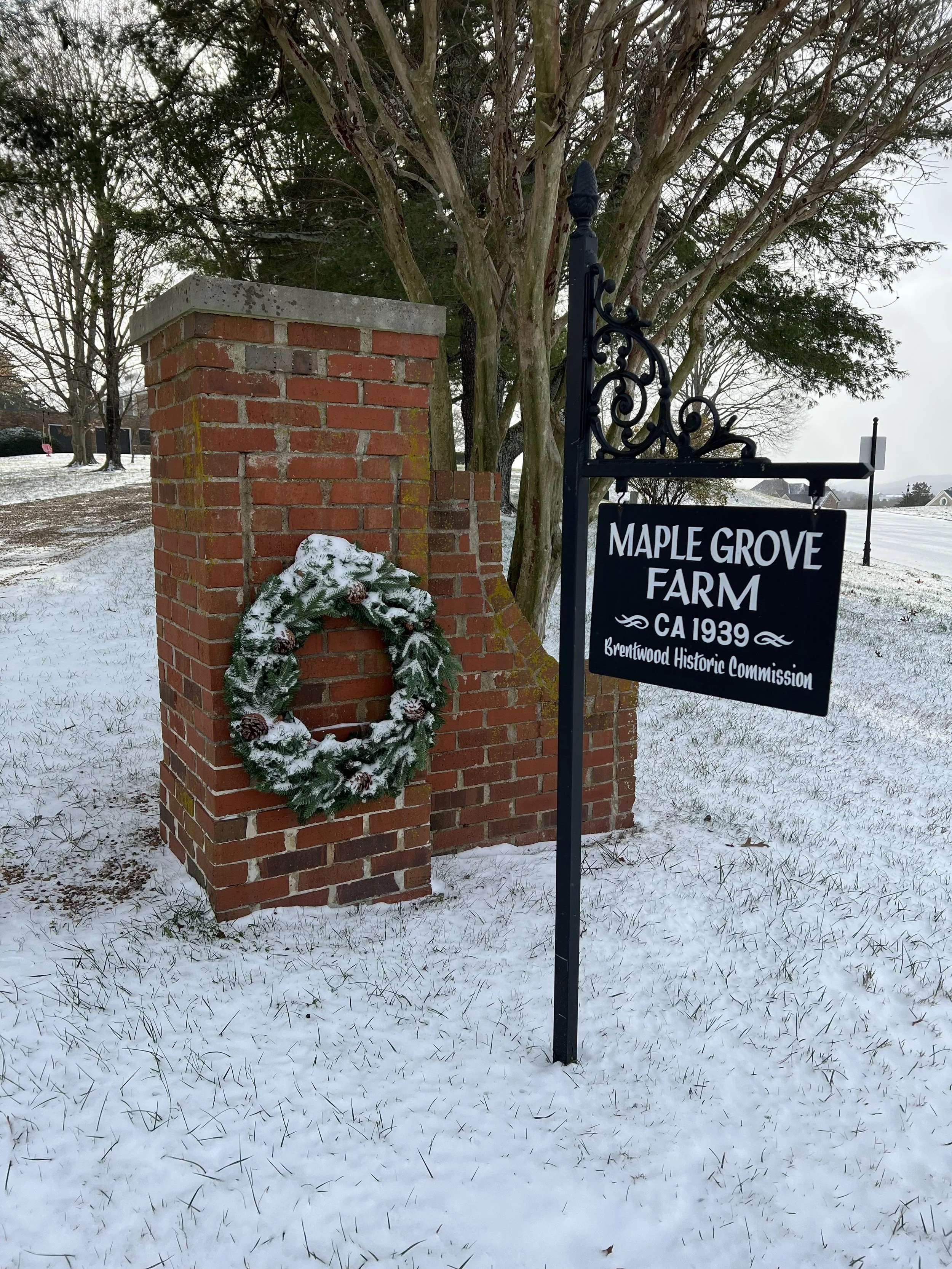 Brick entrance with a decorative wreath, snowy ground, and a black sign that reads "Maple Grove Farm, CA 1939, Brentwood Historic Commission."