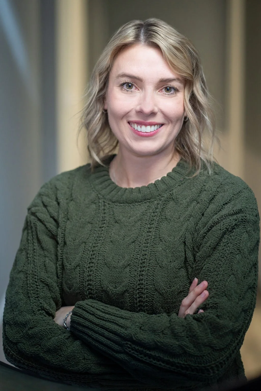 A woman with shoulder-length blonde hair, smiling, wearing a dark green cable-knit sweater, arms crossed.