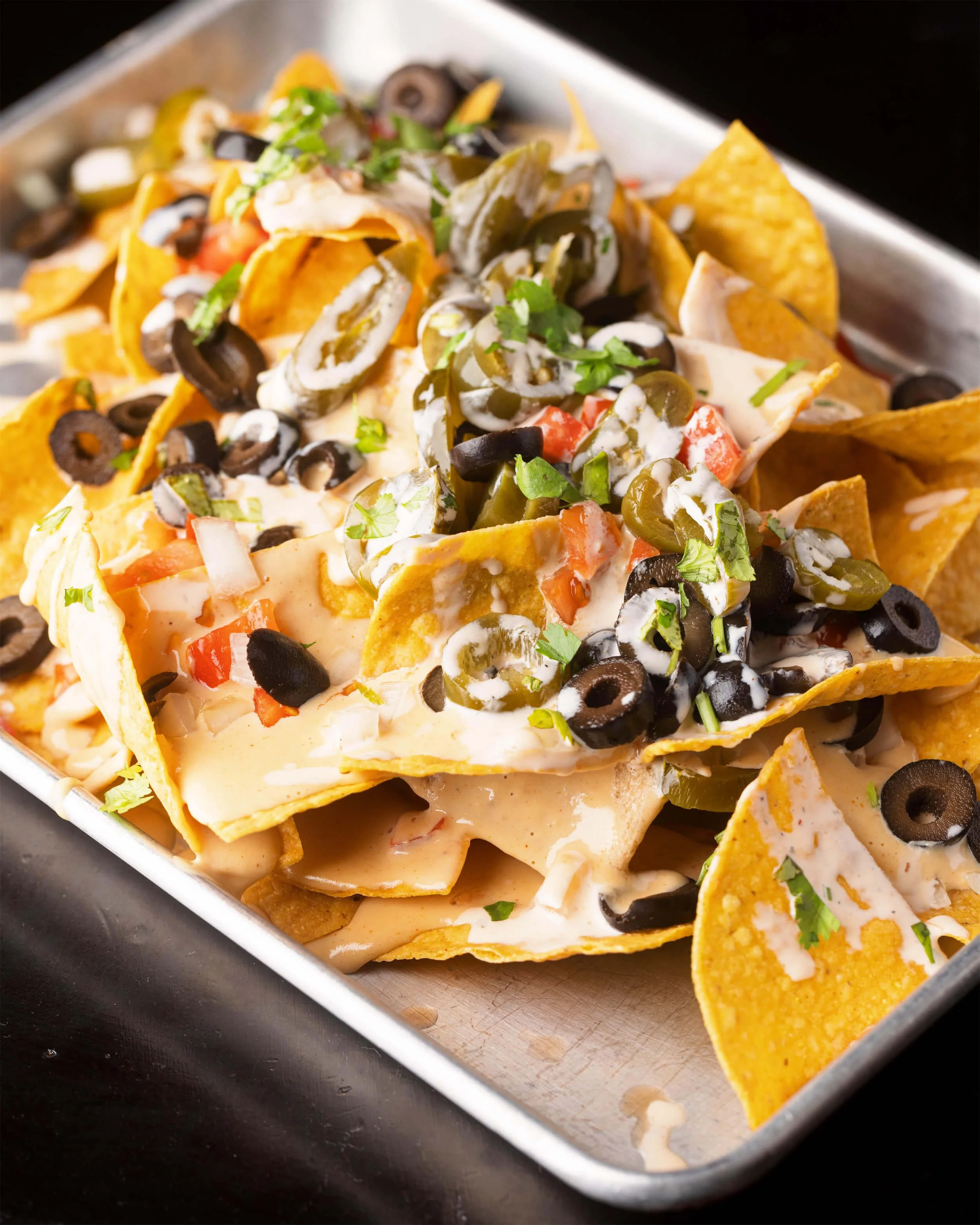 Close-up of a tray of nachos with melted cheese, black and green olives, chopped tomatoes, chopped onions, and fresh cilantro at Ryan's Wake | An Irish Pub, Bar & Restaurant in Troy, NY with River Views, Outdoor Seating & Live Music
