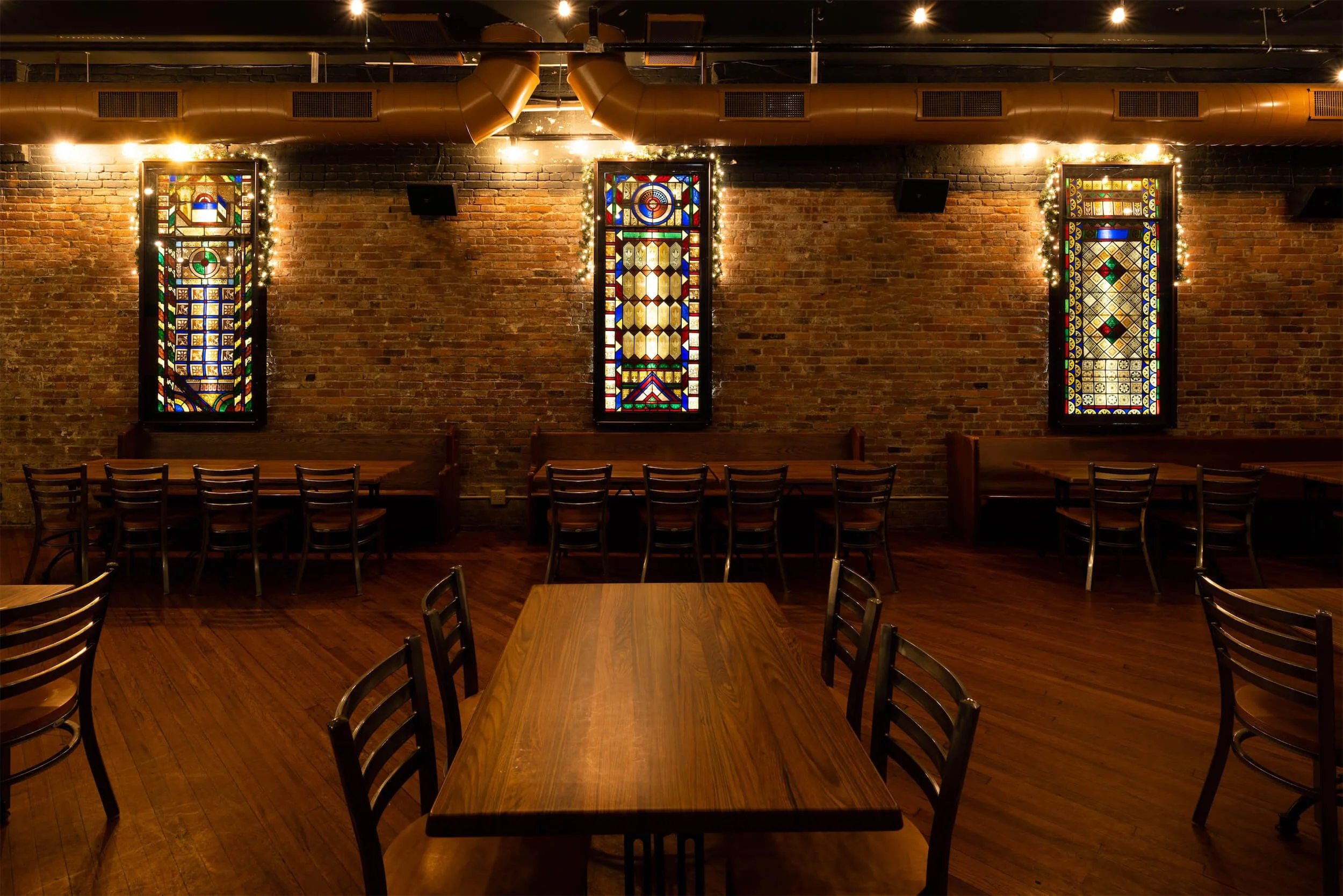 Interior of Ryan's Wake in Troy, NY with wooden tables and chairs, exposed brick wall, stained glass windows illuminated with string lights, and wood-and-metal ceiling ducts.