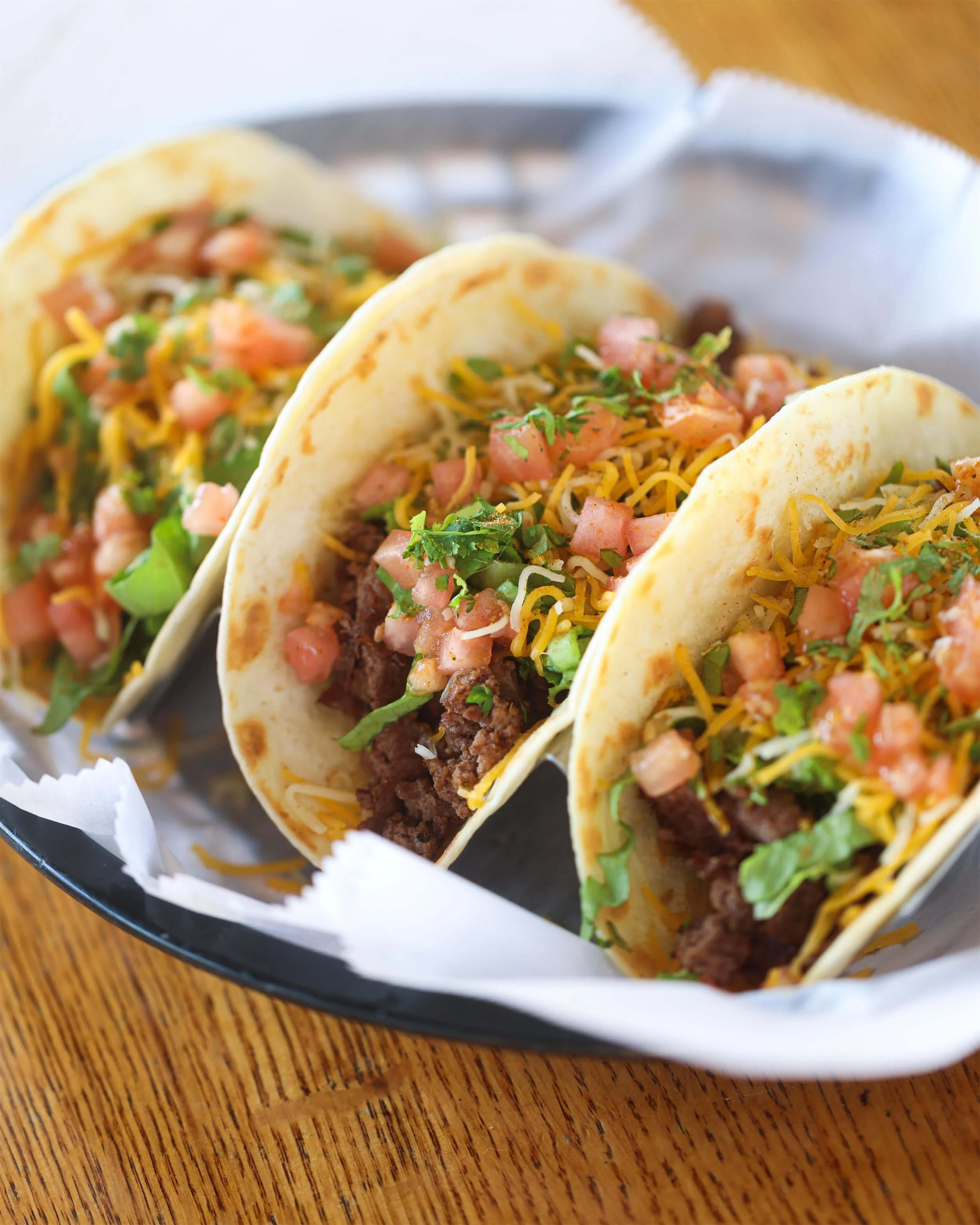Three beef tacos with shredded cheese, diced tomatoes, lettuce, and cilantro in soft corn tortillas on a black plate at Ryan's Wake | An Irish Pub, Bar & Restaurant in Troy, NY with River Views, Outdoor Seating & Live Music