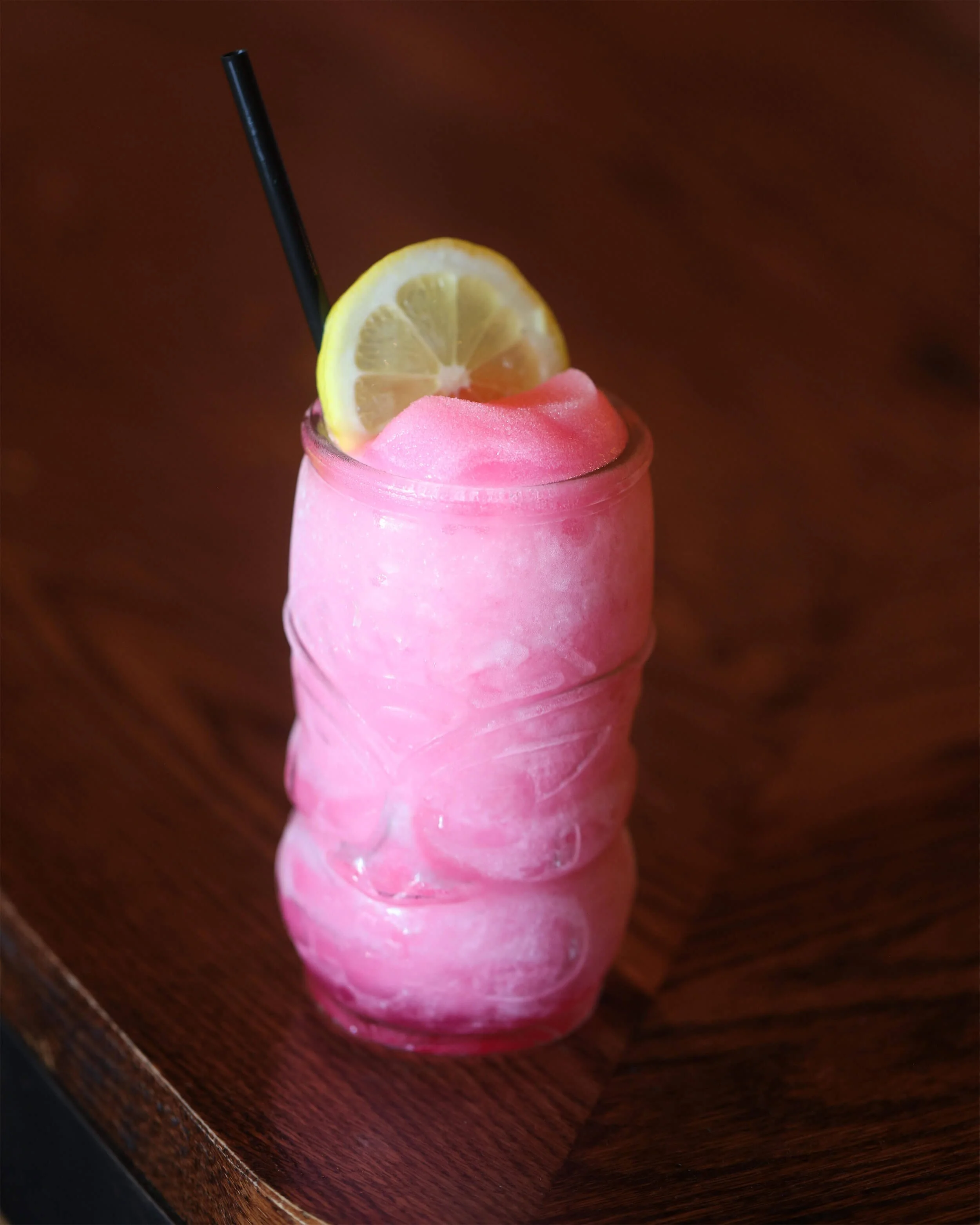 Pink frozen drink in a clear glass with a lemon wedge and a black straw, on a wooden surface  at Ryan's Wake | An Irish Pub, Bar & Restaurant in Troy, NY with River Views, Outdoor Seating & Live Music