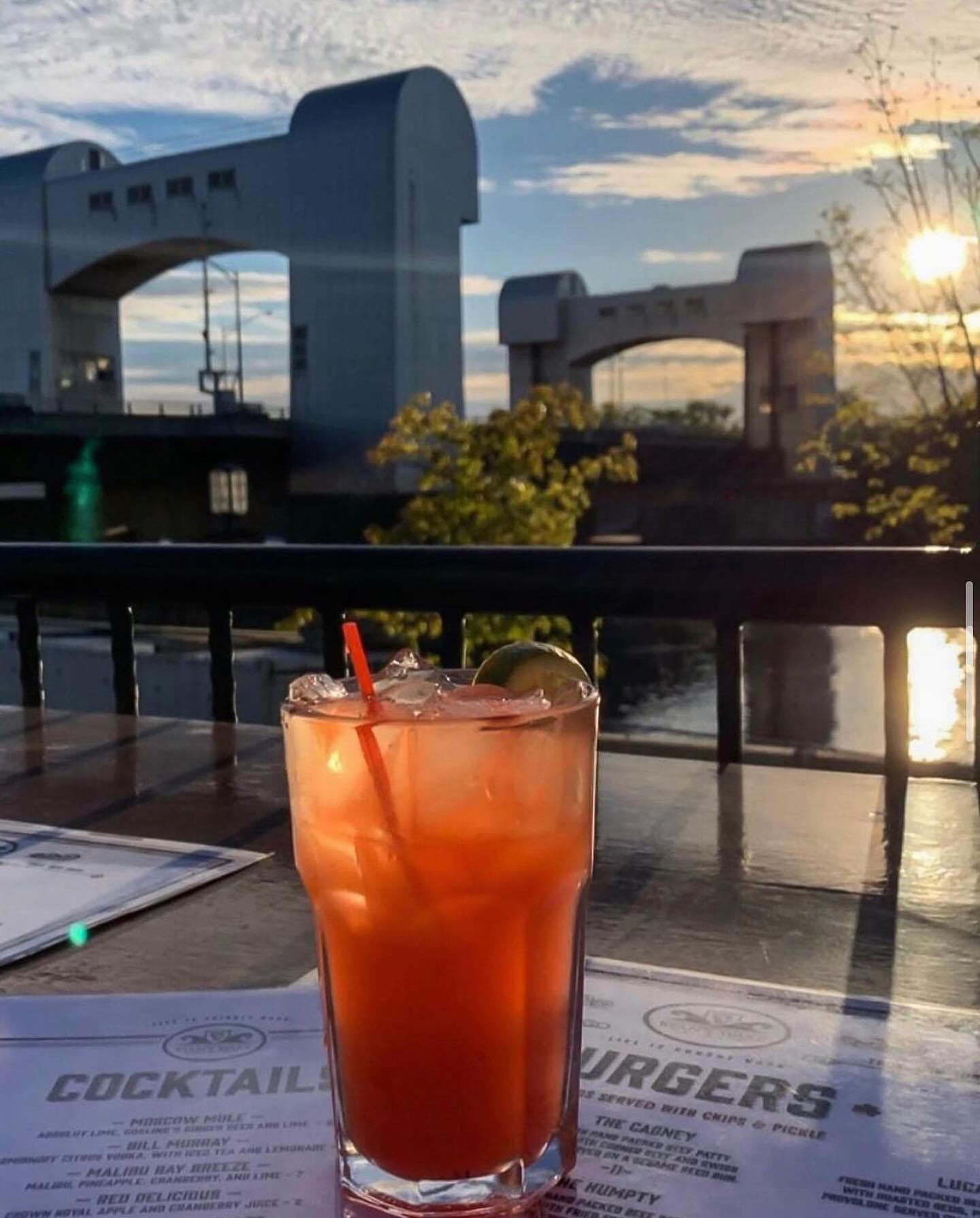 A glass of pink cocktail with ice and lime wedge on a table at outdoor restaurant during sunset, with water and modern bridge structures in the background.